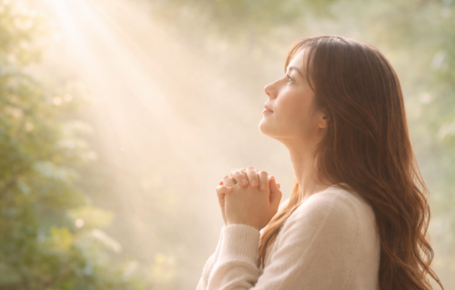 天から差すやわらかな光を見上げ、静かに手を合わせて祈る女性の横顔